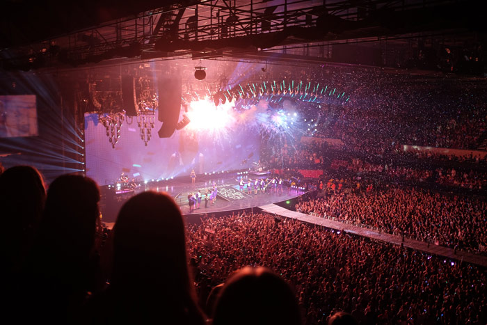 A brightly lit, crowded concert, looking down from the crowd to the stage